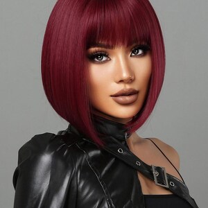 A woman with vibrant red hair styled in a short bob, wearing a black leather jacket and posing against a gray background.