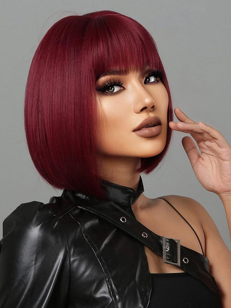 A woman with vibrant red hair styled in a short bob, wearing a black leather jacket and posing with her hand on her chin.