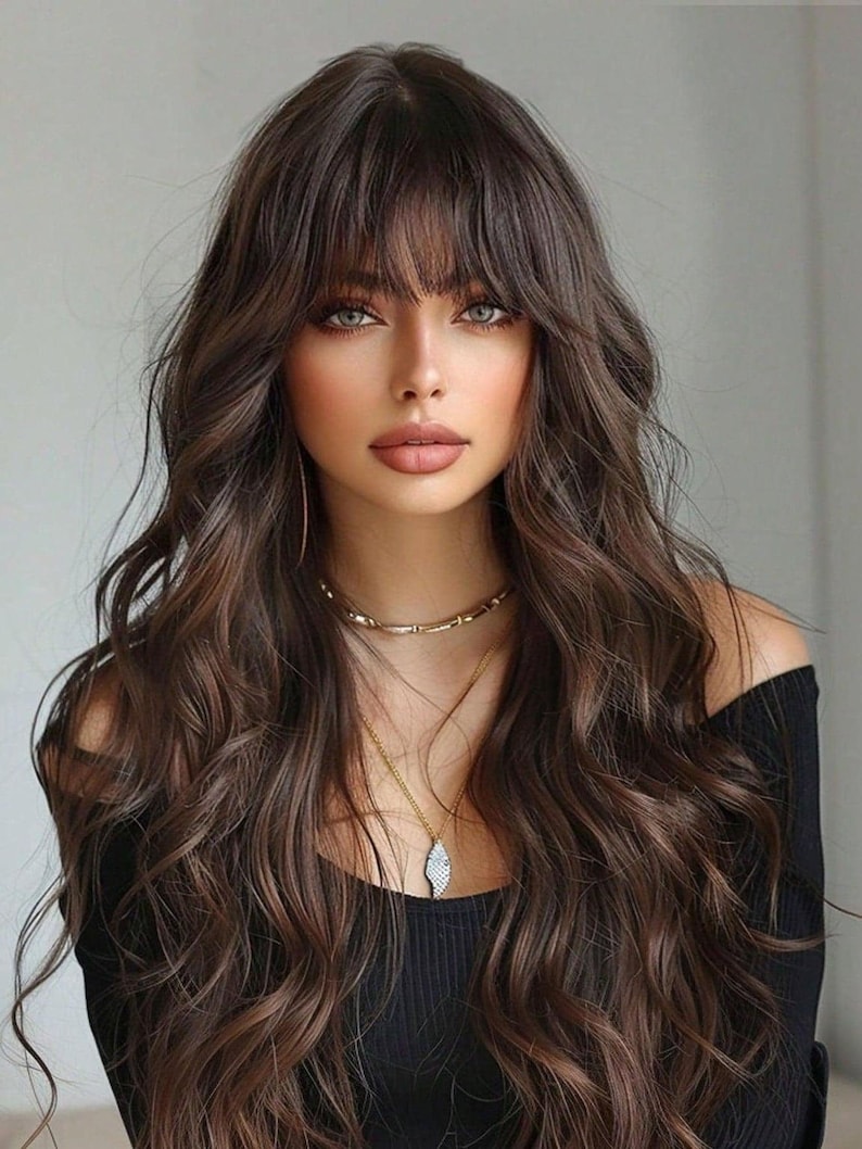 A woman with long, wavy brown hair and bangs, wearing a black top and a necklace, looking directly at the camera with a serious expression.
