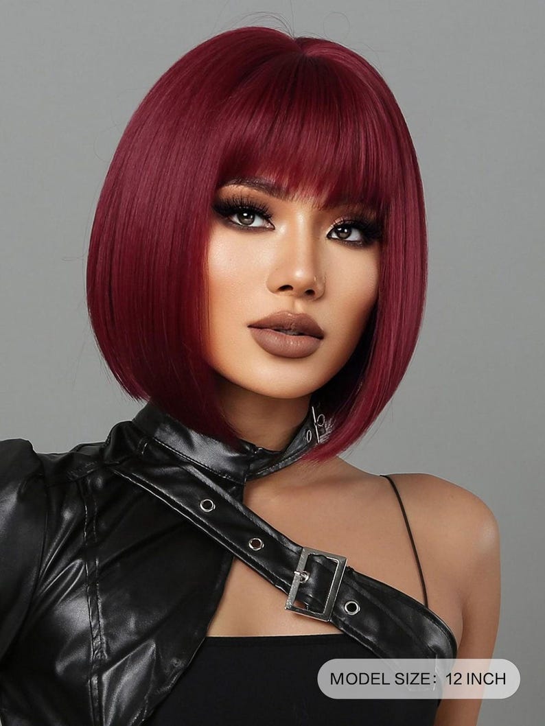 A woman with short, vibrant red hair styled in a bob-like cut, wearing a black leather jacket and posing for a portrait against a gray background.