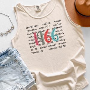 May include: Ivory-colored tank top with the year "1966" printed in colorful letters. The shirt features a list of words associated with the era. A brown hat, denim shorts, and a floral necklace are also in the image.