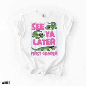 K&ouml;nnte beinhalten: Ein wei&szlig;es T-Shirt mit einem Grafikdruck von drei gr&uuml;nen Alligatoren, die eine schwarze Sonnenbrille tragen. Der Text "SEE YA LATER FIRST GRADER" steht in rosa Buchstaben vor einem Hintergrund aus hellblauen vertikalen Streifen.