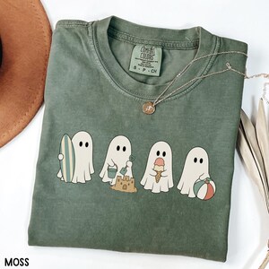 May include: A moss green short-sleeve t-shirt featuring a graphic of four white ghosts engaged in beach activities, including holding a surfboard, building a sandcastle, eating an ice cream cone, and holding a beach ball. The shirt has a crew neck.