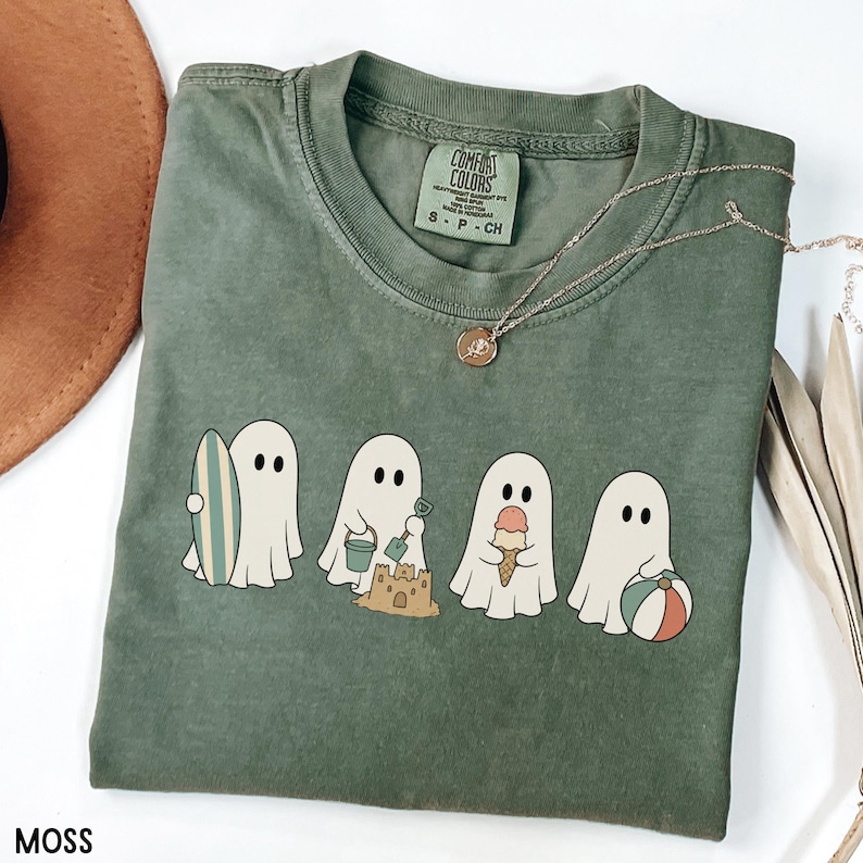 May include: A moss green short-sleeve t-shirt featuring a graphic of four white ghosts engaged in beach activities, including holding a surfboard, building a sandcastle, eating an ice cream cone, and holding a beach ball. The shirt has a crew neck.