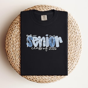 May include: Black t-shirt with the word "Senior" in a blue floral and gingham pattern, with "class of 2026" in white script. The shirt is displayed on a woven, circular surface.