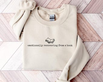 May include: A beige sweatshirt with the text "emotionally recovering from a book" and a small open book graphic in black. The sweatshirt is folded on a wooden surface.