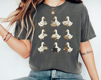 May include: A charcoal gray short-sleeve t-shirt featuring a graphic print of nine white geese in various baking poses, including mixing dough, holding a jar, and carrying a loaf of bread, arranged in a three-by-three grid pattern.