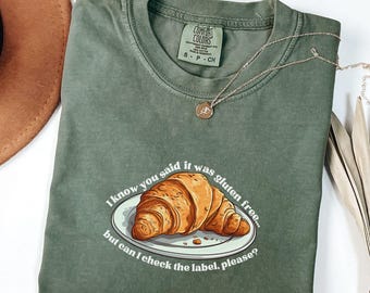 May include: A green t-shirt with a white graphic of a croissant on a plate and the text "I know you said it was gluten free... but can I check the label, please?"