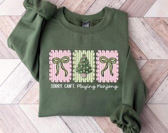 May include: Green sweatshirt with a graphic design featuring pink and green striped rectangles. The design includes a Christmas tree and bows, with the text "SORRY. CAN'T. Playing Mahjong."
