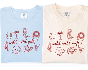 Puede incluir: Dos camisetas, una azul claro y otra marfil, cada una con un gráfico rojo. Los gráficos incluyen una herradura, un cóctel, una bota de vaquero, un lazo, un cactus, naipes, un sombrero de vaquero y una calavera de toro. La camiseta azul claro dice "wild wild gals" y la camiseta marfil dice "wild wild wife".