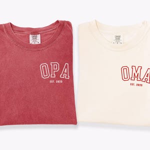 May include: Two folded t-shirts, one red and one cream-colored. The red shirt has the word "OPA" and "EST. 2025" printed in white. The cream shirt has the word "OMA" and "EST. 2025" printed in red. Both shirts have the "Comfort Colors" logo.