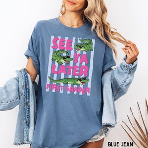 K&ouml;nnte beinhalten: Ein blaues T-Shirt mit Rundhalsausschnitt und einer Grafik von drei gr&uuml;nen Alligatoren mit schwarzer Sonnenbrille. Der Text "SEE YA LATER FIRST GRADER" steht in rosa Buchstaben auf einem Hintergrund aus hellblauen und wei&szlig;en Streifen.