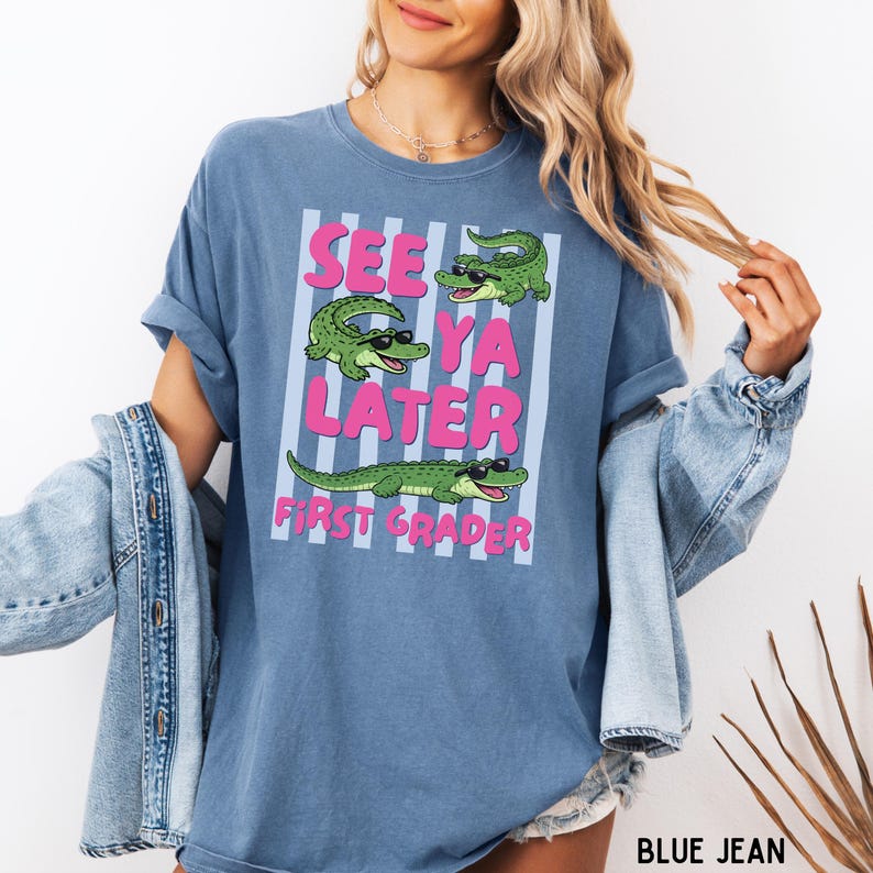 K&ouml;nnte beinhalten: Ein blaues T-Shirt mit Rundhalsausschnitt und einer Grafik von drei gr&uuml;nen Alligatoren mit schwarzer Sonnenbrille. Der Text "SEE YA LATER FIRST GRADER" steht in rosa Buchstaben auf einem Hintergrund aus hellblauen und wei&szlig;en Streifen.