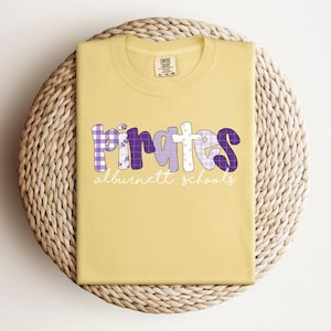 May include: A yellow t-shirt with the word "Pirates" in purple and white patchwork letters, with "alburnett schools" in white script underneath. The shirt is folded on a woven, circular surface.