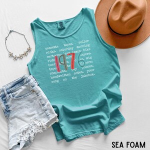 K&ouml;nnte beinhalten: T&uuml;rkisfarbenes Tanktop mit dem Text "1976" und einer Wortliste. Das Oberteil wird mit Jeansshorts, einer Halskette und einem braunen Hut kombiniert. Das Shirt hat einen Rundhalsausschnitt und ist aus weichem Material.