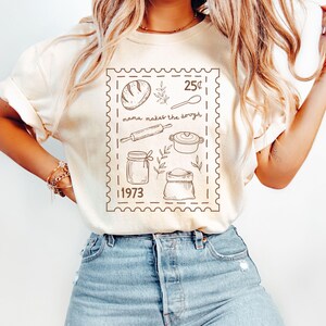 May include: A beige t-shirt with a brown graphic of a vintage postage stamp. The stamp features a loaf of bread, a rolling pin, a jar, a bag of flour, a pot, and a spoon. The text on the stamp reads "25¢ mama makes the dough 1973".