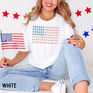 May include: White t-shirt with a graphic of the American flag made of hot dogs and drinks. The word "WHITE" is printed at the bottom of the image. The person is holding a small American flag.