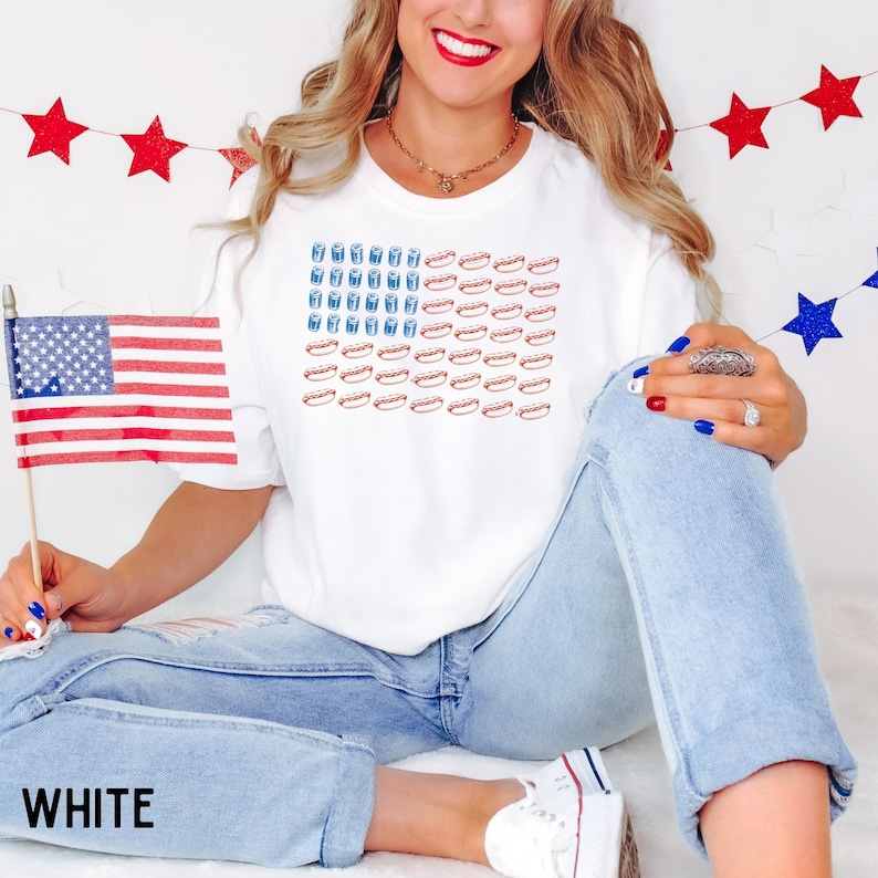 May include: White t-shirt with a graphic of the American flag made of hot dogs and drinks. The word "WHITE" is printed at the bottom of the image. The person is holding a small American flag.