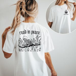 Puede incluir: Camiseta blanca con un gr&aacute;fico negro en la espalda que dice "read in peace". El gr&aacute;fico presenta un esqueleto leyendo un libro en un campo de flores y hierba. Una versi&oacute;n m&aacute;s peque&ntilde;a del gr&aacute;fico est&aacute; en la parte delantera.