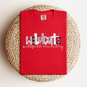 May include: A red t-shirt with the word "Wildcats" in a patchwork design, featuring black and white patterns, and gray hearts. Below, the text "smithfield elementary" is written in a cursive font. The shirt is displayed on a woven, circular surface.