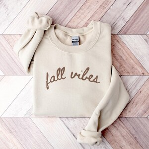May include: A beige sweatshirt with the words "fall vibes" in brown script. The long sleeves are cuffed, and the sweatshirt is folded neatly. The background is a light brown and white wooden pattern.