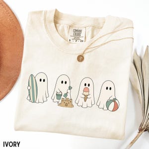 May include: An ivory t-shirt featuring a graphic of four white ghosts engaged in summer activities. The ghosts are shown holding a surfboard, building a sandcastle with a bucket and shovel, eating an ice cream cone, and holding a beach ball.