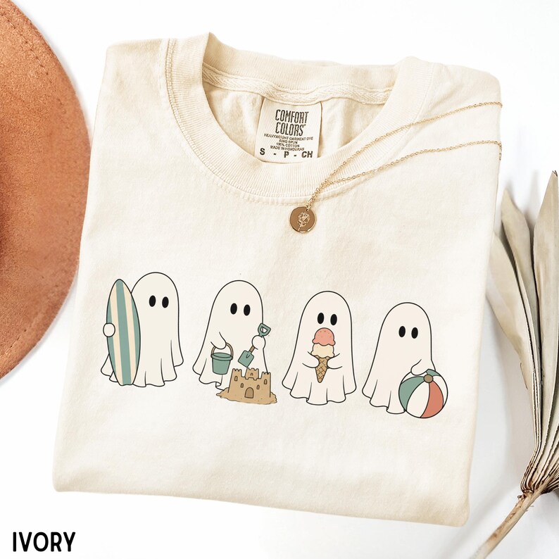 May include: An ivory t-shirt featuring a graphic of four white ghosts engaged in summer activities. The ghosts are shown holding a surfboard, building a sandcastle with a bucket and shovel, eating an ice cream cone, and holding a beach ball.