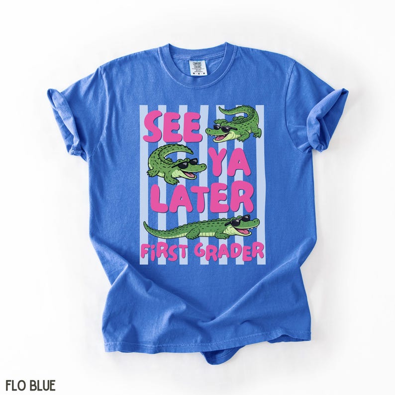 K&ouml;nnte beinhalten: Ein blaues T-Shirt mit einer Grafik von drei gr&uuml;nen Krokodilen, die schwarze Sonnenbrillen tragen. Der Text "SEE YA LATER FIRST GRADER" ist in rosa Buchstaben gedruckt. Das Design liegt auf einem Hintergrund mit vertikalen wei&szlig;en Streifen.