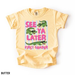 K&ouml;nnte beinhalten: Ein buttergelbes T-Shirt mit einer Grafik von drei gr&uuml;nen Alligatoren, die eine schwarze Sonnenbrille tragen. Der Text "SEE YA LATER FIRST GRADER" steht in rosa Buchstaben auf hellblauen vertikalen Streifen. Das T-Shirt hat einen Rundhalsausschnitt.