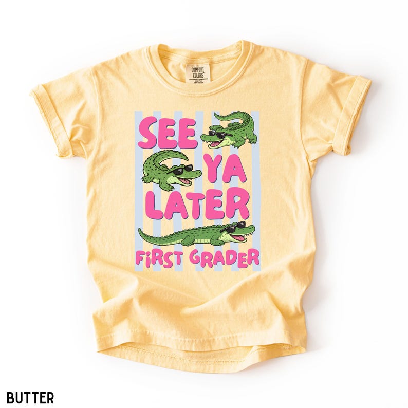 K&ouml;nnte beinhalten: Ein buttergelbes T-Shirt mit einer Grafik von drei gr&uuml;nen Alligatoren, die eine schwarze Sonnenbrille tragen. Der Text "SEE YA LATER FIRST GRADER" steht in rosa Buchstaben auf hellblauen vertikalen Streifen. Das T-Shirt hat einen Rundhalsausschnitt.