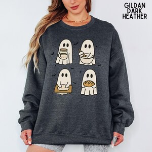 May include: A dark heather gray crewneck sweatshirt featuring a graphic of four white ghosts baking. One ghost holds a jar, one stirs a bowl, one kneads dough on a board, and one holds a loaf of bread. Small black bats are scattered around the ghosts.