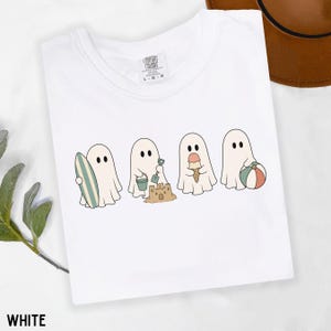 May include: A white t-shirt featuring a graphic of four ghosts engaged in beach activities. The ghosts are shown holding a surfboard, building a sandcastle with a bucket and shovel, eating an ice cream cone, and holding a colorful beach ball.