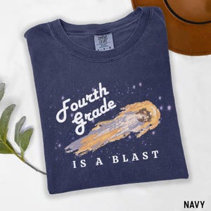 May include: A navy blue short-sleeve t-shirt with a graphic print of a comet and the text Fourth Grade IS A BLAST. The shirt features a crew neckline and a Comfort Colors label on the inside collar indicating a size large.