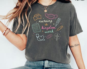 May include: A charcoal gray short-sleeve t-shirt featuring the text "nursing is kingdom work" in colorful script and block lettering. The design includes medical icons like a stethoscope, syringe, thermometer, bandage, clipboard, and a cross.