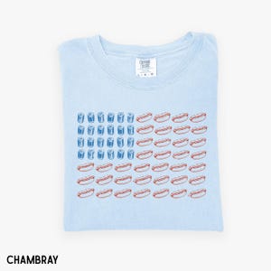 May include: Light blue t-shirt with a graphic design of an American flag made of hot dogs and blue cylinders. The shirt is folded and has a tag that reads "Comfort Colors". The word "Chambray" is printed at the bottom.