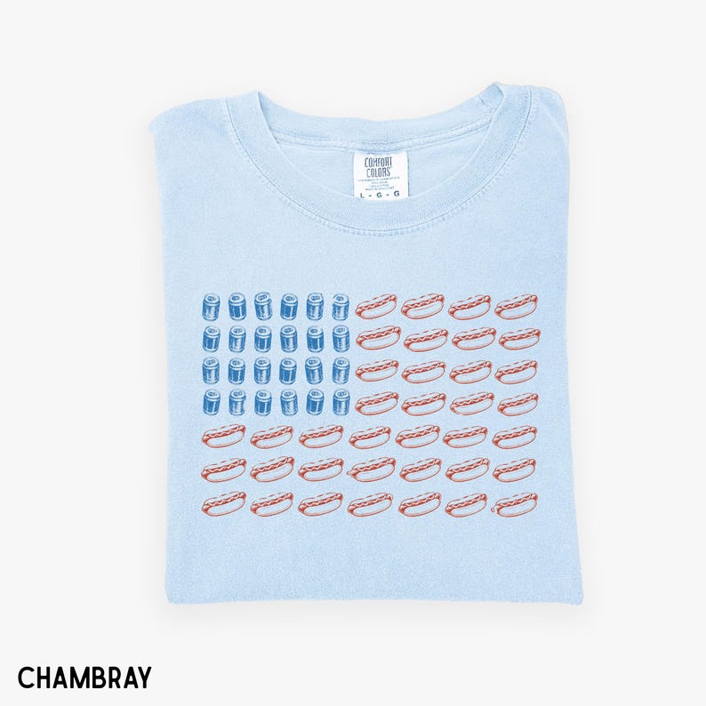 May include: Light blue t-shirt with a graphic design of an American flag made of hot dogs and blue cylinders. The shirt is folded and has a tag that reads "Comfort Colors". The word "Chambray" is printed at the bottom.