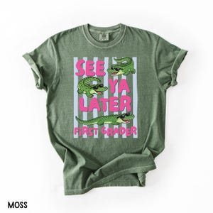 K&ouml;nnte beinhalten: Ein moosgr&uuml;nes Kurzarm-T-Shirt mit einer Grafik von drei gr&uuml;nen Alligatoren, die schwarze Sonnenbrillen tragen. Der Text "SEE YA LATER FIRST GRADER" steht in fetten rosa Buchstaben auf einem Hintergrund aus hellblauen und wei&szlig;en Streifen.