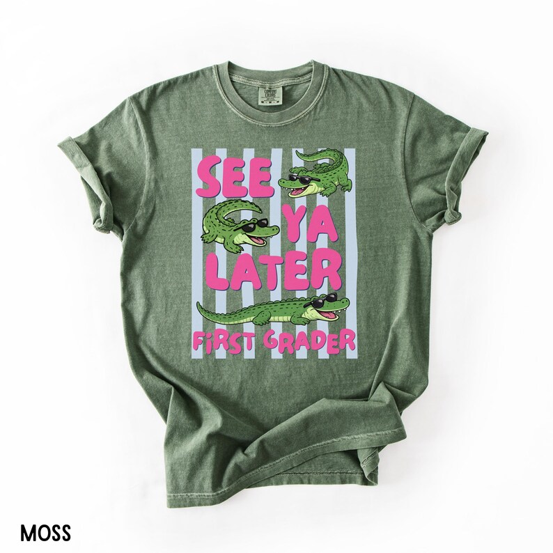K&ouml;nnte beinhalten: Ein moosgr&uuml;nes Kurzarm-T-Shirt mit einer Grafik von drei gr&uuml;nen Alligatoren, die schwarze Sonnenbrillen tragen. Der Text "SEE YA LATER FIRST GRADER" steht in fetten rosa Buchstaben auf einem Hintergrund aus hellblauen und wei&szlig;en Streifen.