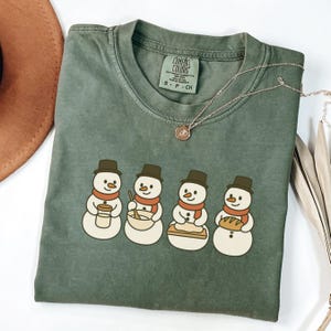 May include: An olive green Comfort Colors t-shirt featuring a graphic of four snowmen. Each snowman wears a black hat and a red scarf, and holds a different item: a coffee cup, a mixing bowl, a pie, and a loaf of bread. A gold necklace is draped across the shirt.