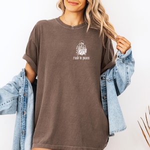 Puede incluir: Camiseta marr&oacute;n con mangas enrolladas. Presenta un gr&aacute;fico blanco de un esqueleto leyendo un libro, con el texto "read in peace" debajo. La camiseta tiene un corte holgado y un aspecto vintage.