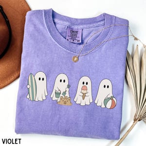 May include: A violet short-sleeved t-shirt featuring a graphic of four white ghosts engaged in beach activities, including holding a surfboard, building a sandcastle with a bucket and shovel, eating an ice cream cone, and holding a beach ball.