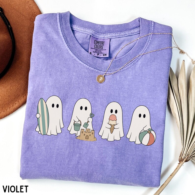 May include: A violet short-sleeved t-shirt featuring a graphic of four white ghosts engaged in beach activities, including holding a surfboard, building a sandcastle with a bucket and shovel, eating an ice cream cone, and holding a beach ball.
