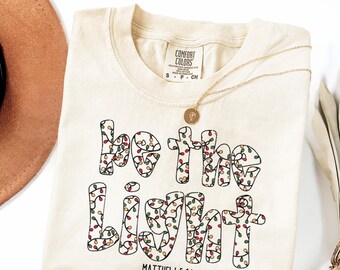 May include: Cream-colored t-shirt with the phrase "Be the Light" in a festive font, decorated with colorful string lights. The text "Matthew 5:14" is below the phrase. A gold necklace and a brown hat are also in the image.