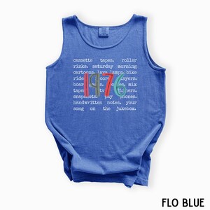 K&ouml;nnte beinhalten: Ein blaues Tanktop mit dem Jahr "1976" in farbigem Text. Das Shirt zeigt eine Liste nostalgischer Gegenst&auml;nde und Phrasen aus den 1970er Jahren, darunter Kassetten, Lavalampe und Samstagmorgen-Cartoons. Der Text "FLO BLUE" steht unten.