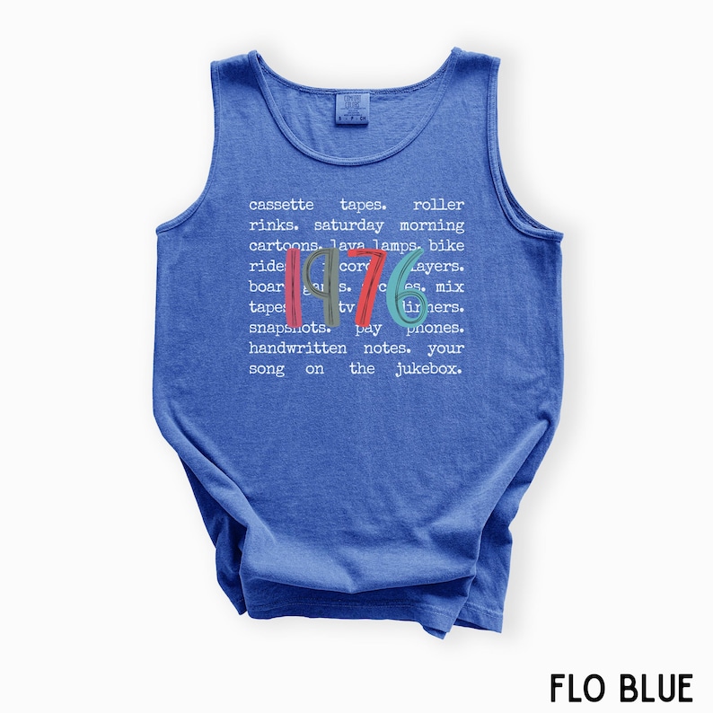 K&ouml;nnte beinhalten: Ein blaues Tanktop mit dem Jahr "1976" in farbigem Text. Das Shirt zeigt eine Liste nostalgischer Gegenst&auml;nde und Phrasen aus den 1970er Jahren, darunter Kassetten, Lavalampe und Samstagmorgen-Cartoons. Der Text "FLO BLUE" steht unten.