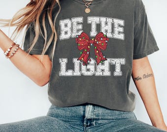 May include: Dark gray t-shirt with the text "BE THE LIGHT" in white block letters. A red bow with Christmas lights is centered between the words. The shirt is a casual style, suitable for everyday wear.