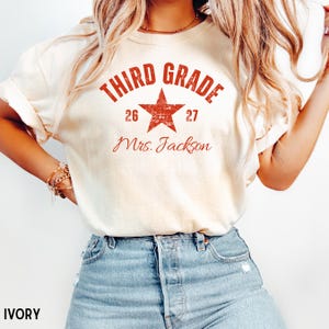 May include: An ivory short-sleeved t-shirt featuring a distressed red graphic with the text "THIRD GRADE" arched over a star, the numbers "26" and "27" on either side, and the name "Mrs. Jackson" in a cursive script font below the star.