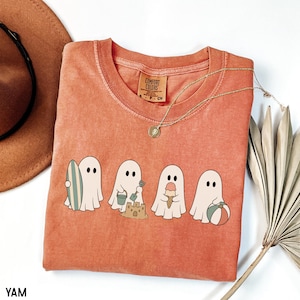 May include: A terracotta-colored short-sleeve t-shirt featuring a graphic of four white ghosts engaged in beach activities. One ghost holds a surfboard, one builds a sandcastle with a bucket and shovel, one holds an ice cream cone, and one holds a beach ball.