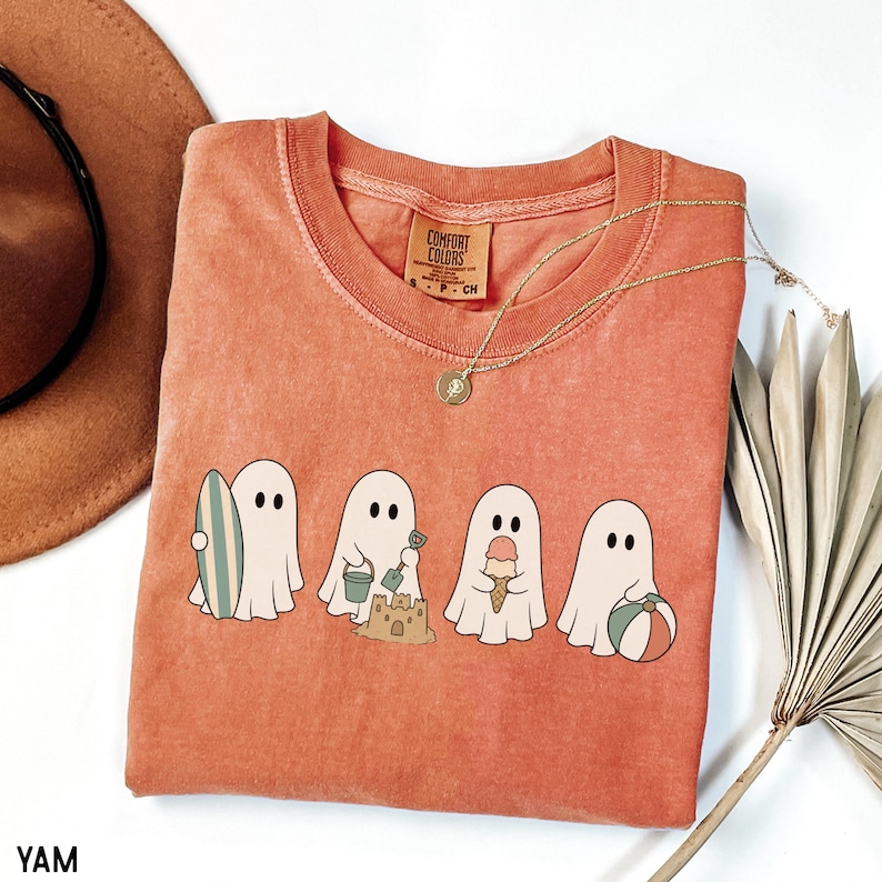 May include: A terracotta-colored short-sleeve t-shirt featuring a graphic of four white ghosts engaged in beach activities. One ghost holds a surfboard, one builds a sandcastle with a bucket and shovel, one holds an ice cream cone, and one holds a beach ball.