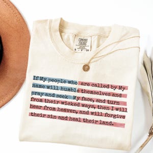 May include: Cream-colored t-shirt with the text "If My people who are called by My name will humble themselves and pray and seek My face..." in black. A gold necklace with a pendant rests on the shirt. A brown hat is partially visible.